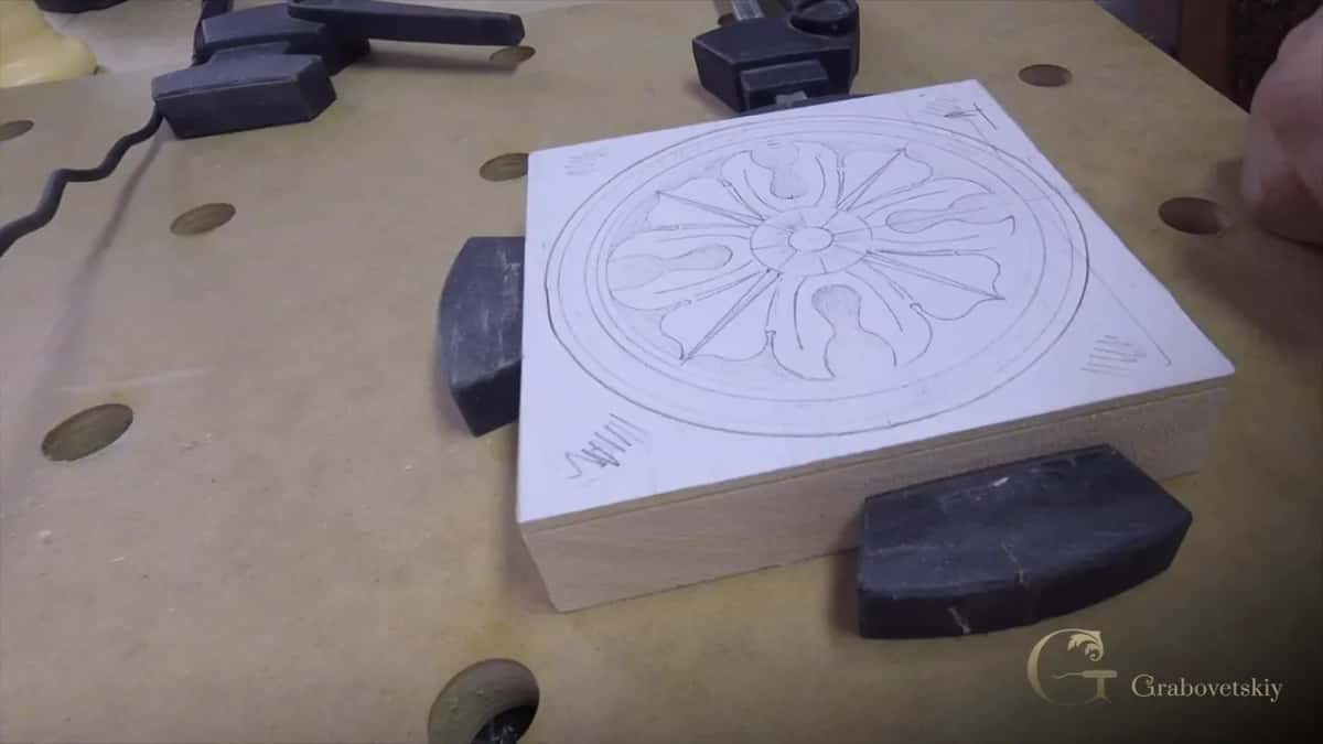 A square blank held by bench dogs. The rosette is finished in pencil, no wood is off yet, and the block edge shows the fibre.