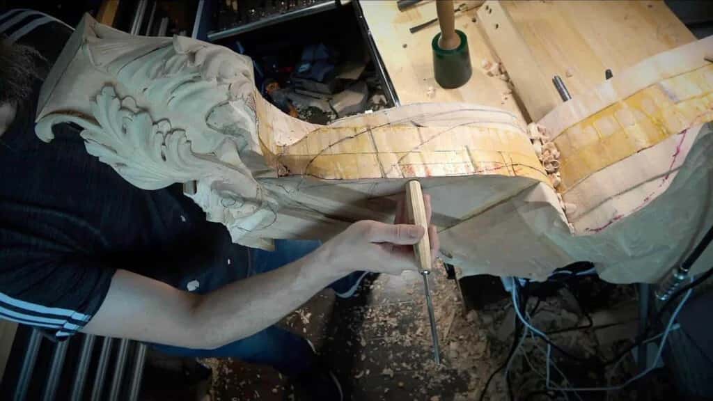 Hands working a large carved scroll, the piece propped up on trestles in the yard