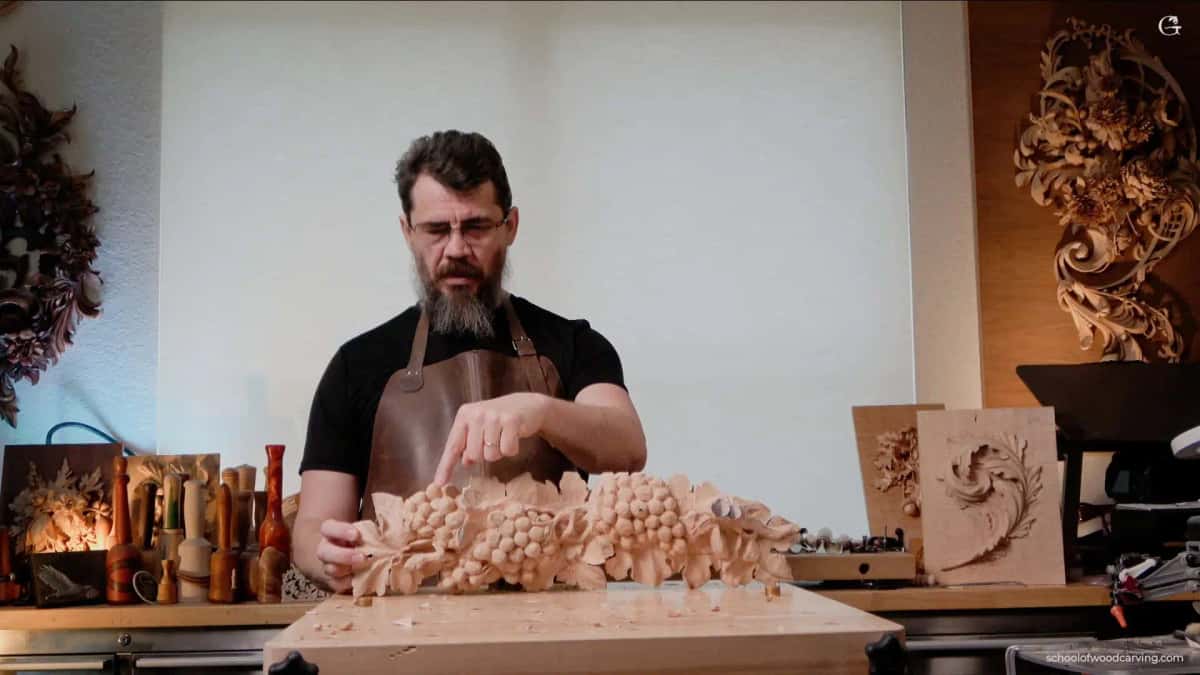 Alexander Grabovetskiy holds a finished grape garland onlay across his bench in a leather apron, with carved panels and turned mallets filling the shelves behind him