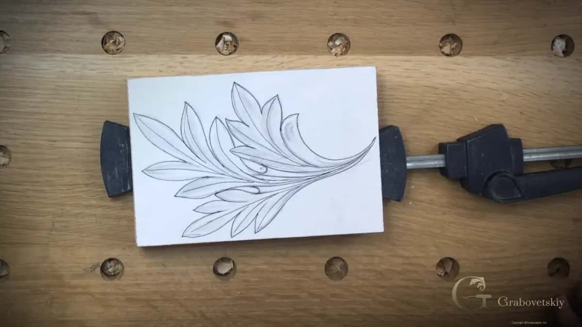 Pencil drawing of an acanthus leaf on a white faced blank, pinched between two clamps on a dog hole bench