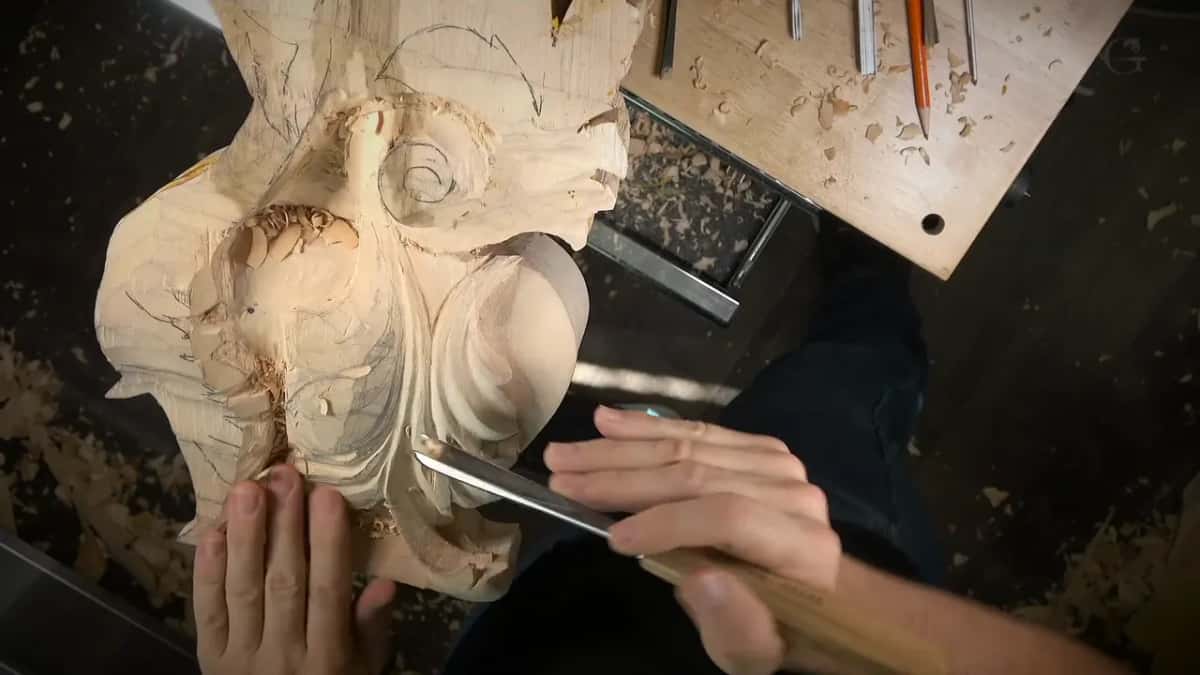 A long gouge driven into the edge of a pale roughed leaf relief, the other hand holding the panel steady.