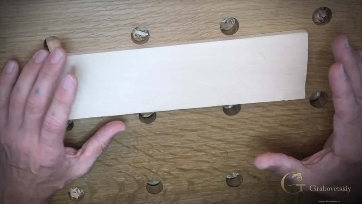 Both hands flatten a pale basswood plank on an oak bench drilled with dog holes.