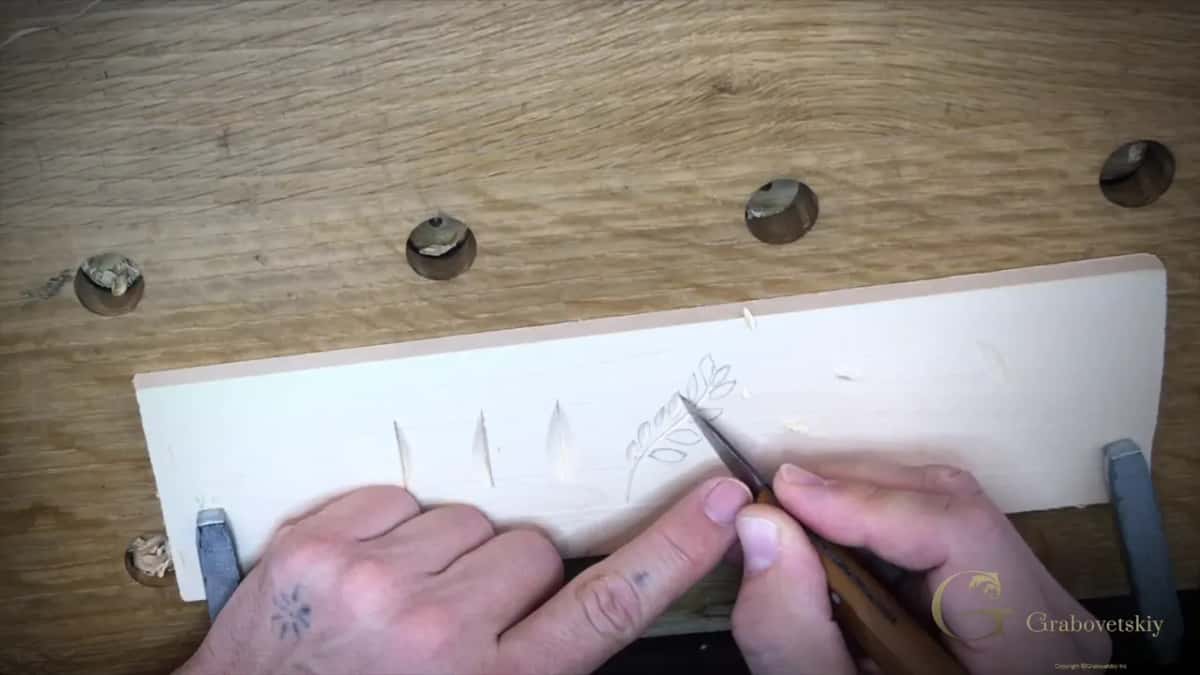 Three practice nicks and one carved leaf sprig on a pale board, the knife tip still in the wood.