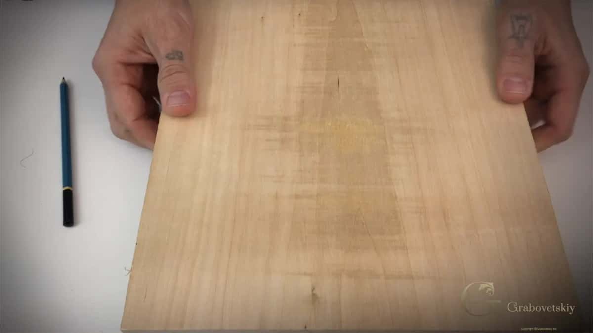 Hands hold a pale board with faint grain against a white top, a sharpened pencil resting beside it.