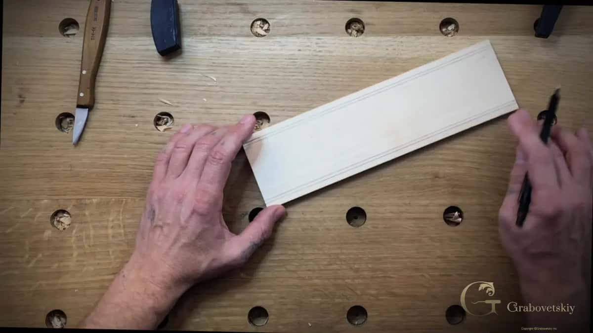A pale board ruled with two pencil border lines waits beside a carving knife and a chisel.