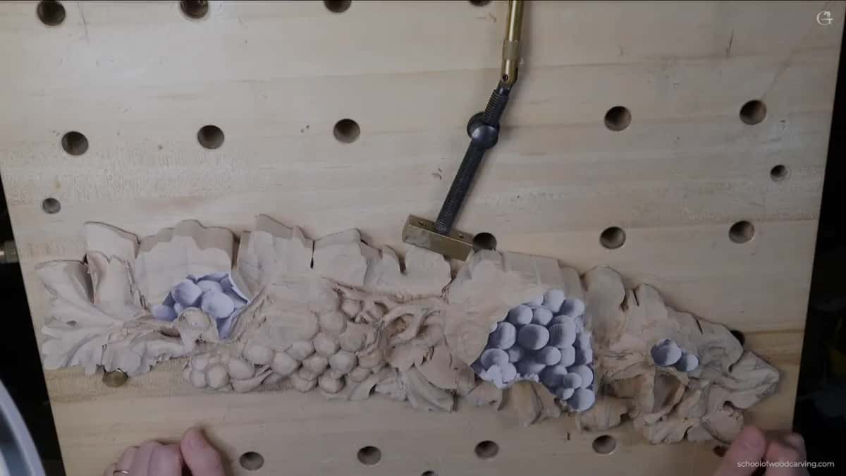 A pale panel of grapes and leaves, half roughed out, clamped to a pale bench.