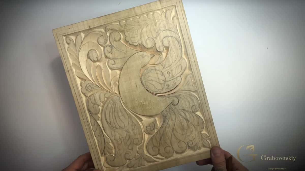 Both hands lift a pale panel where a bird and leaves are half carved and half still pencil.