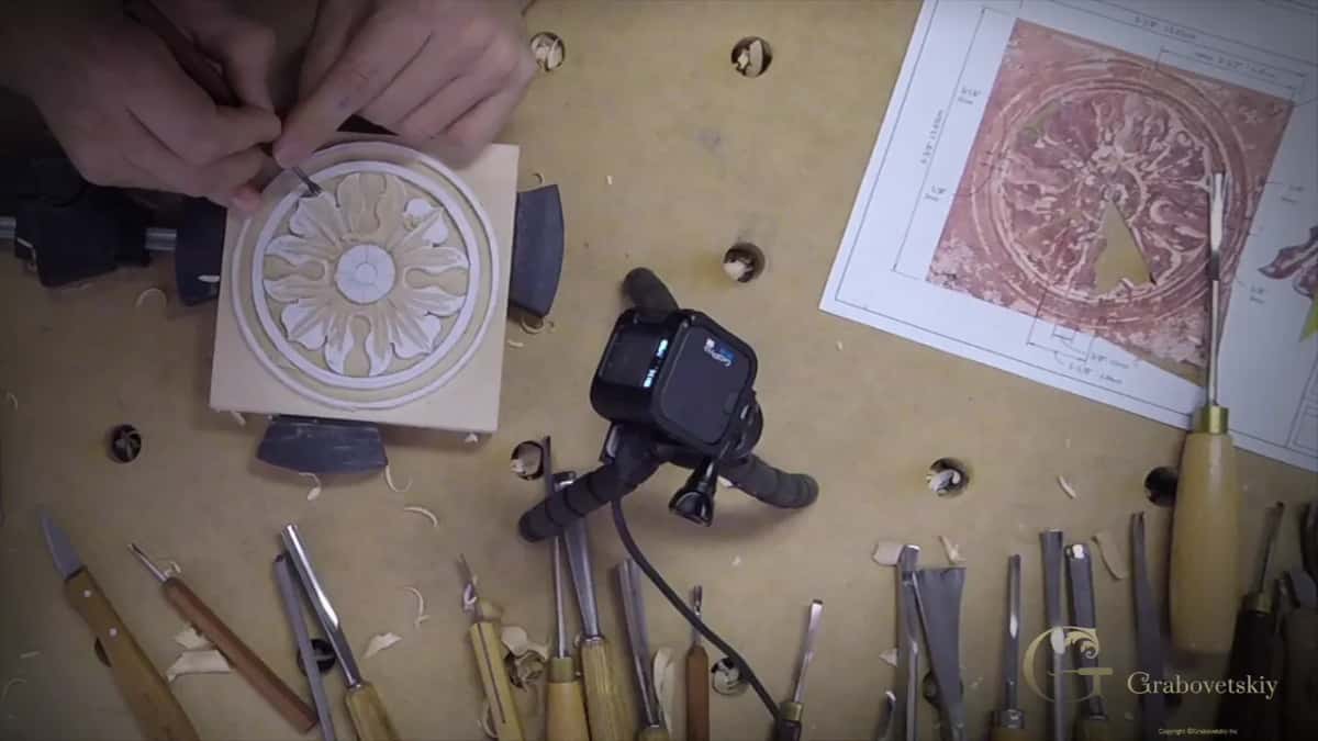 Hands carve a small pale rosette while fifteen gouges and knives lie in a row along the bench.