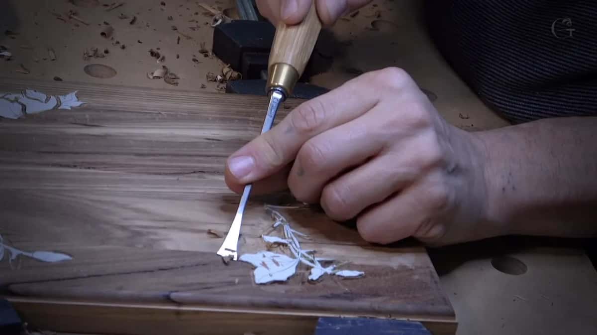 A V-tool carves a small leaf into a dark board of strong stripes, the fresh cuts showing pale.