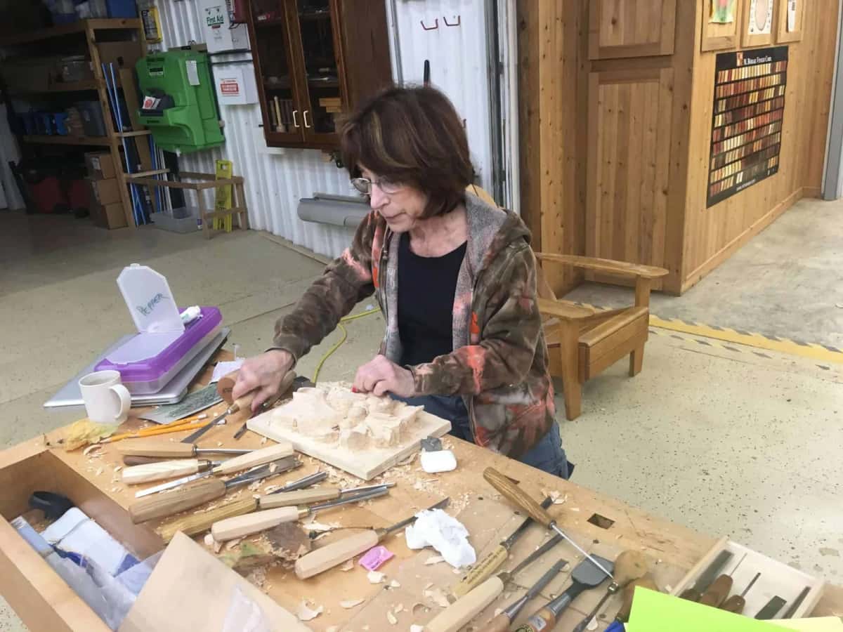 A woman carving a relief panel at her own bench, coffee going cold behind the tool roll