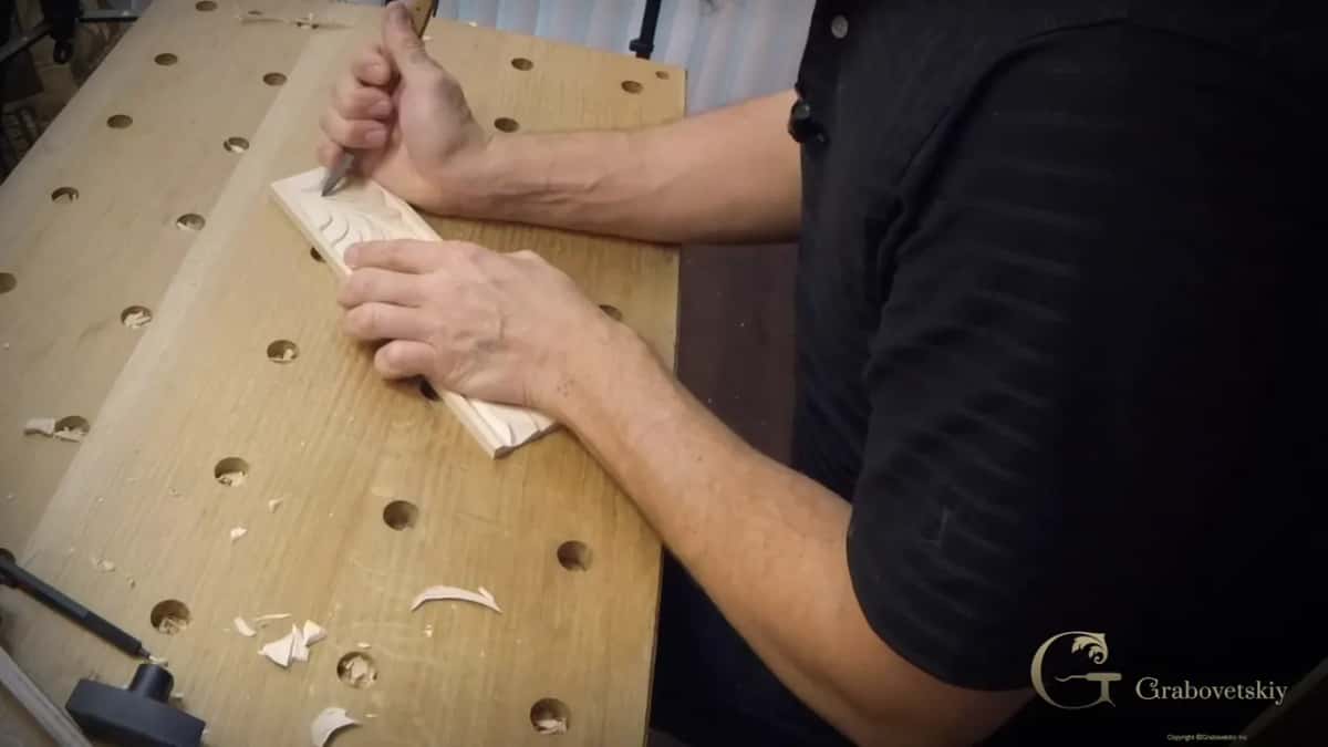 Knife carving a narrow basswood strip with fresh chips scattered across the bench around the work