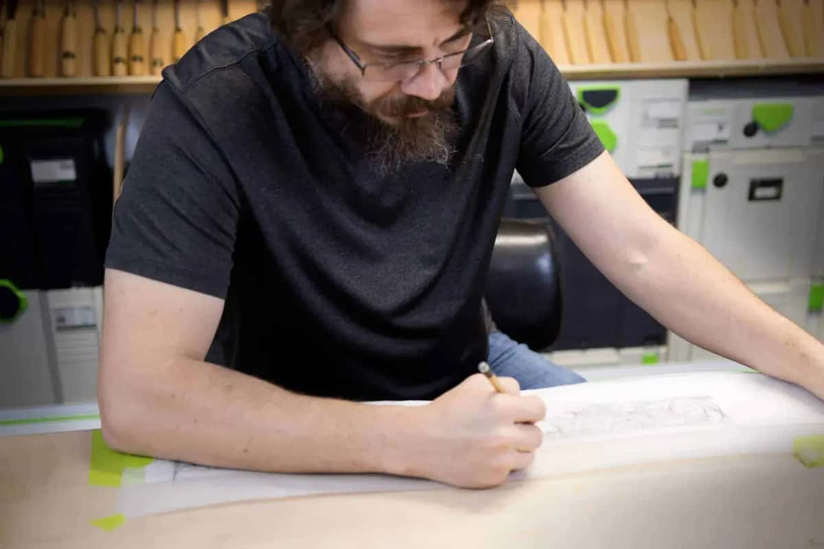 Alexander Grabovetskiy drawing a furniture ornament in pencil at the bench, carving gouges racked on the wall behind him