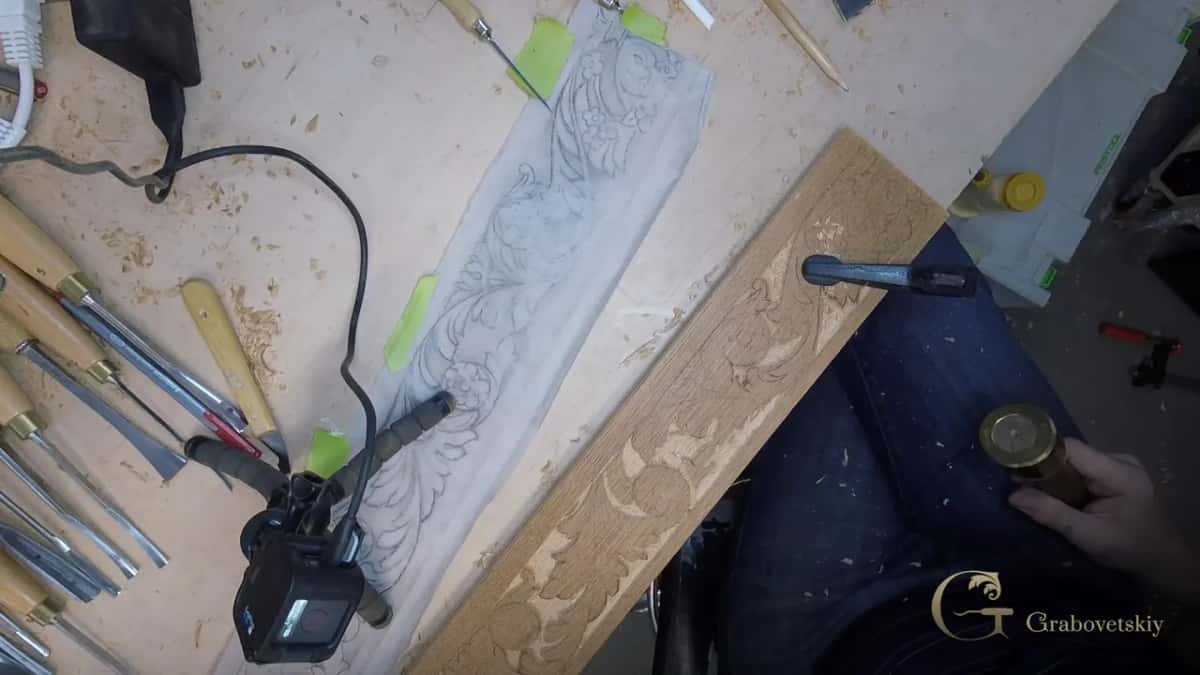 Looking down on the bench, a carved border along a tan board, a row of gouges laid out, brass mallet in his hand.