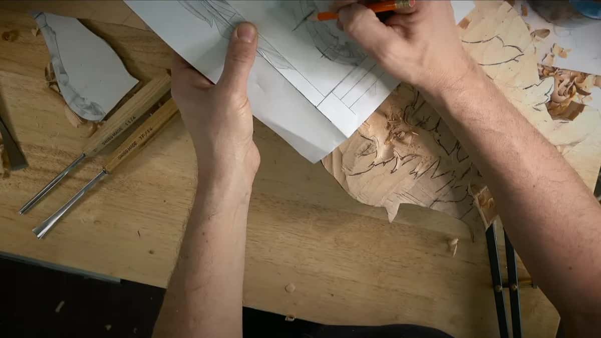Both hands hold a printed drawing over the block while an orange pencil marks a line on the paper