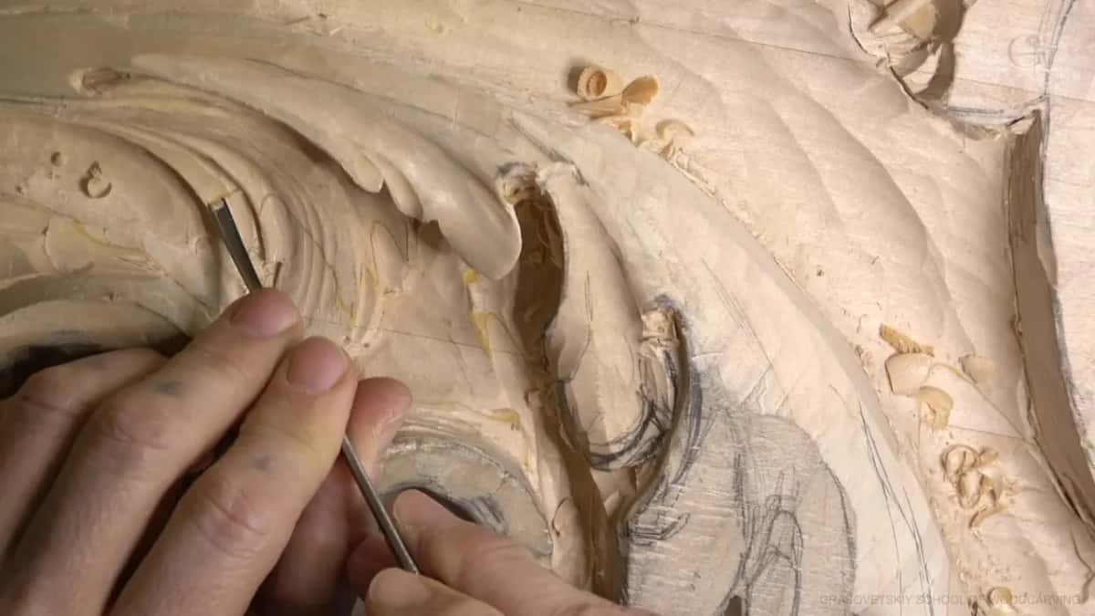 Slim tool angled down into one lobe, steadied by fingers, with chips scattered on the carved surface nearby.