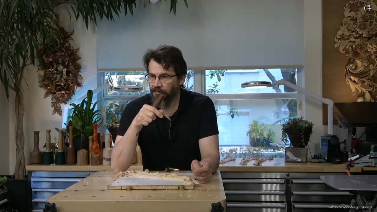 Alexander holding a tool up at his bench, carved panels on the wall behind him