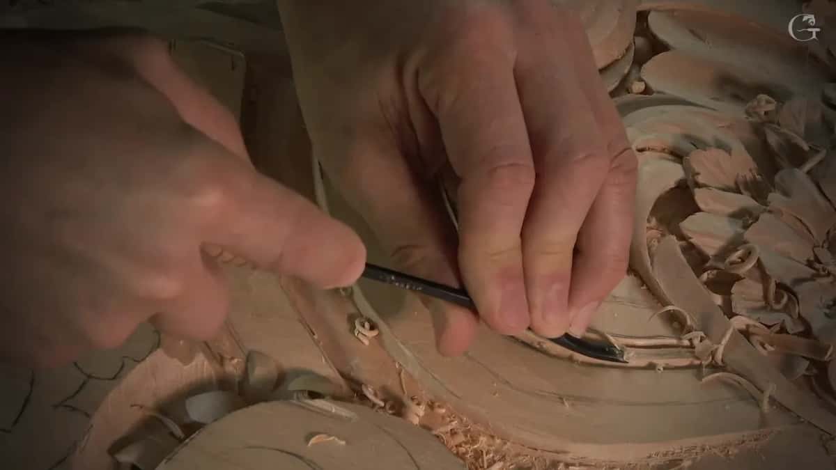 Two fingers pinch a bent tool whose tip sits down in the groove below the carved leaves