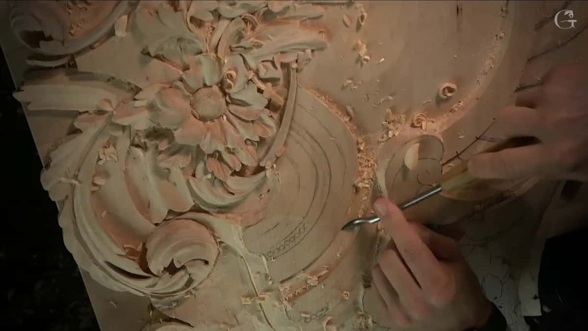 Both hands work a bent tool along the band, and above them sits the carved flower