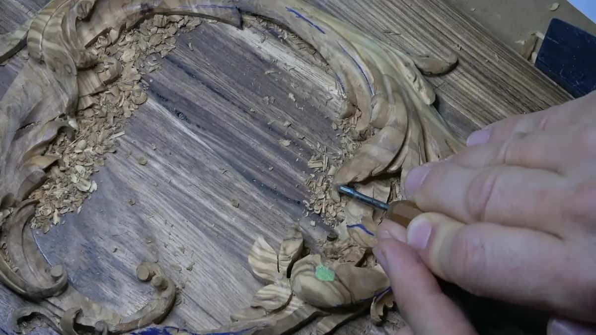 Olive acanthus curling around a dark acacia lid, chips inside the ring, a hand pushing a short gouge into the leaves