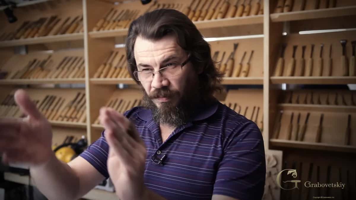 Alexander explaining which way grain wants to be carved, standing in front of the rack he built, gouges behind him.