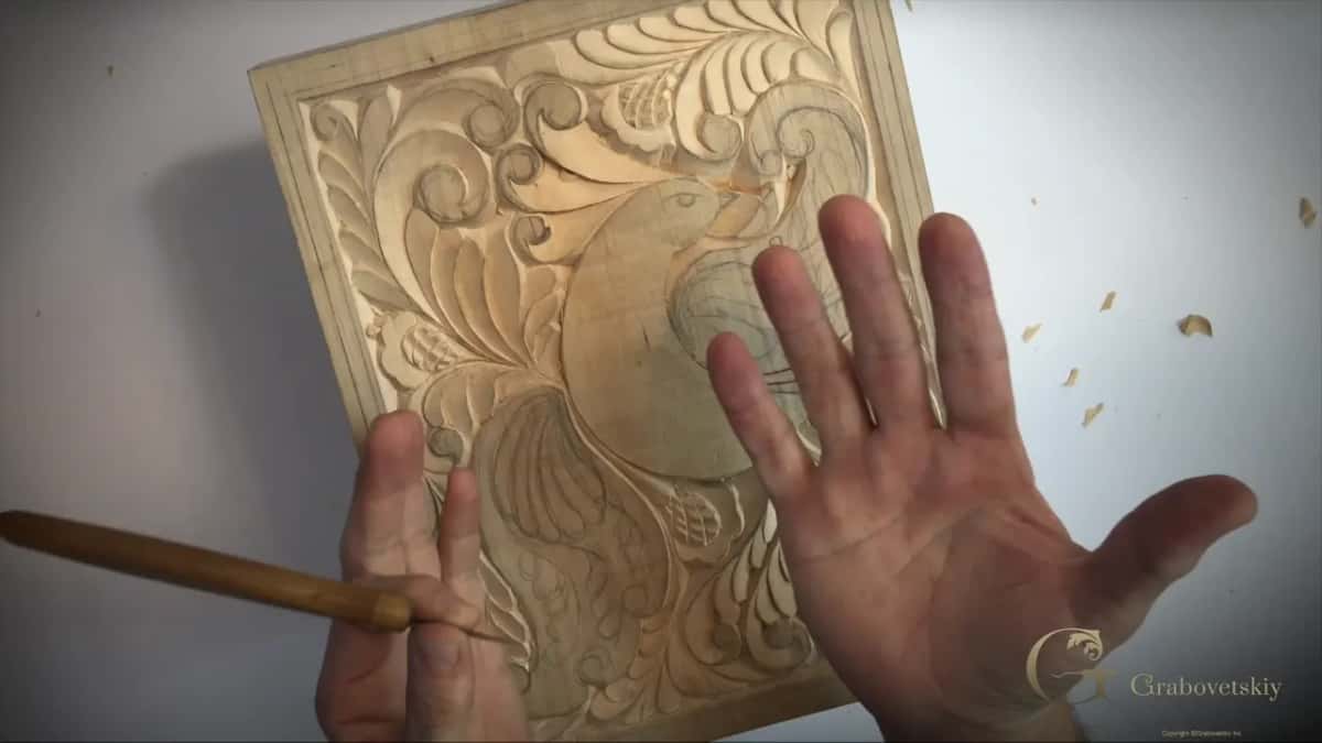 Both hands resting on the half carved board, the knife still held in one of them, chips scattered across the bench beside it.