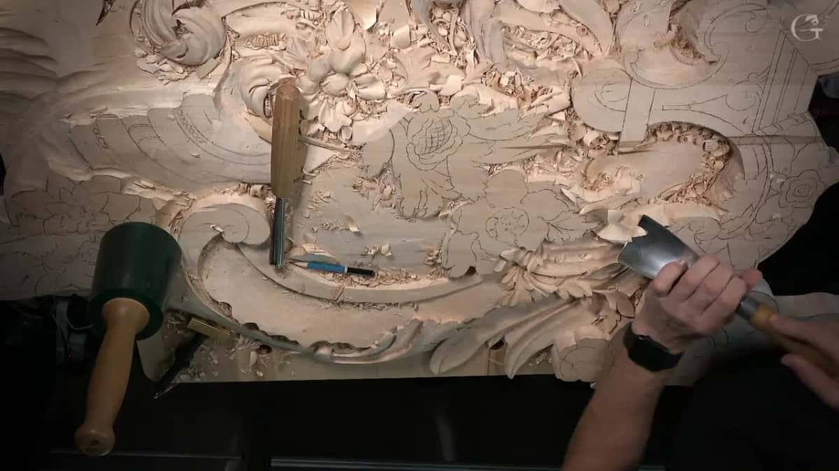 From overhead, a wide gouge in both hands at the right edge, other tools resting on the bench