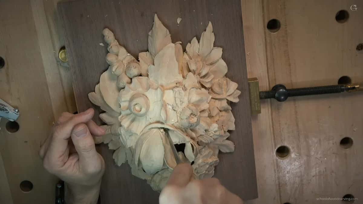 Overhead of the half finished panel clamped down on a dark board, with a small gouge opening a hole beneath one petal
