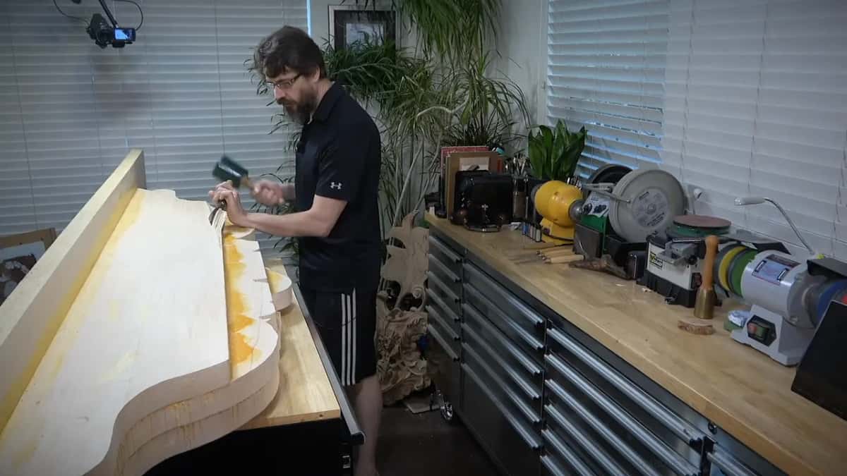 Standing at a long pale block, a mallet in one hand and a big gouge in the other