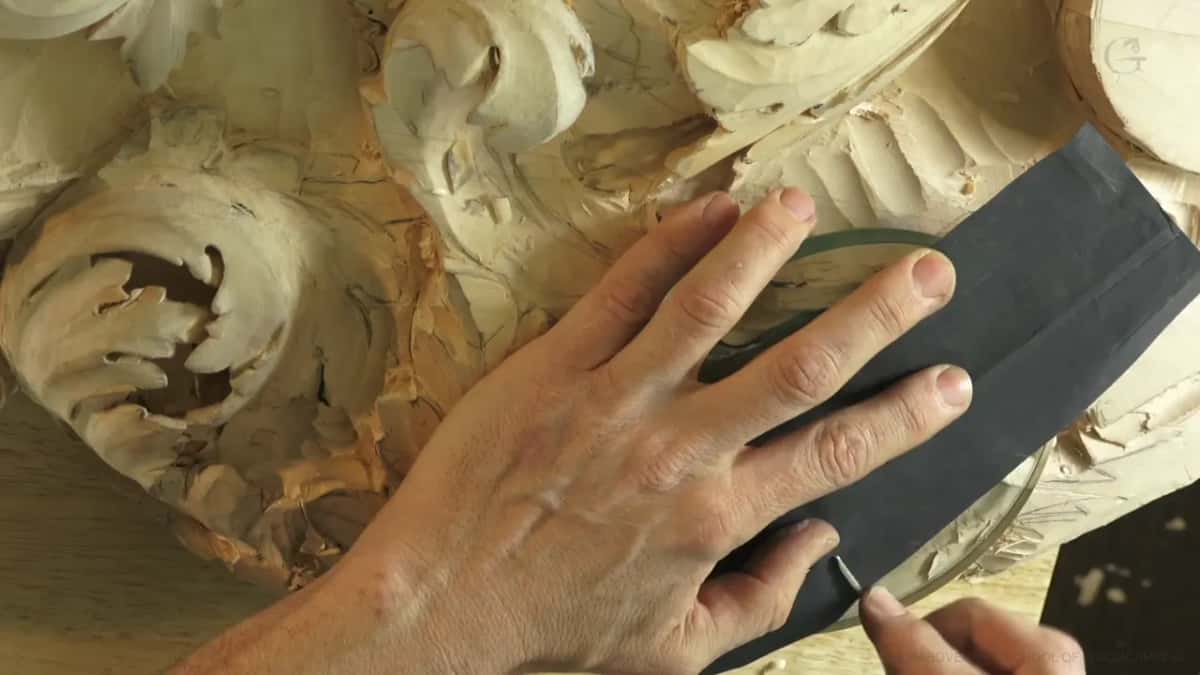 One hand holding black sandpaper flat on a sheet of glass beside a carved acanthus