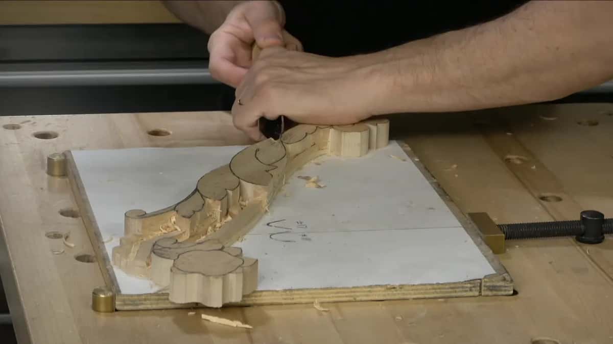 Both hands guiding a tool along the edge of a scrolled molding clamped to a white board on his bench