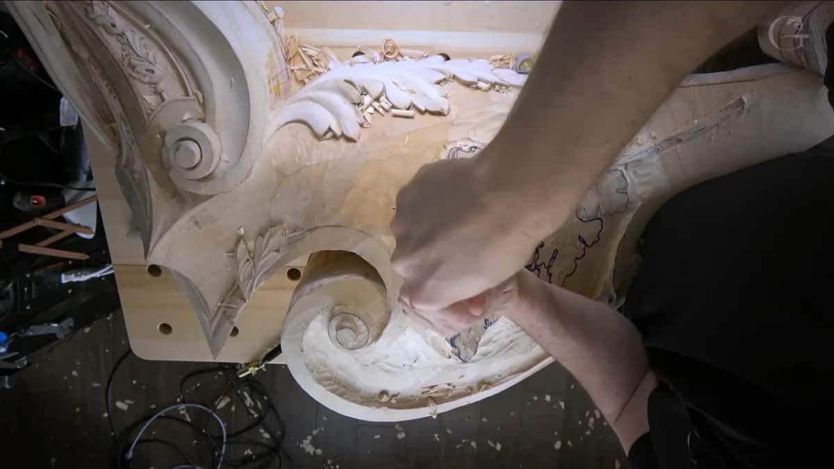 Hands braced on a gouge at the lower oak leaf, the carved acanthus and volute just beside them