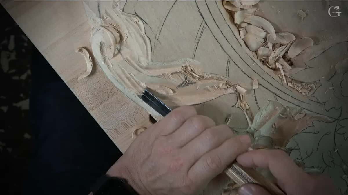 Gouge held low at the leaf while a curl of wood lies to the left of it on the panel