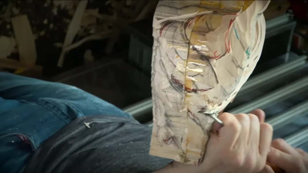Seen from above, a knee in blue jeans under the block and one hand steering a gouge near its base