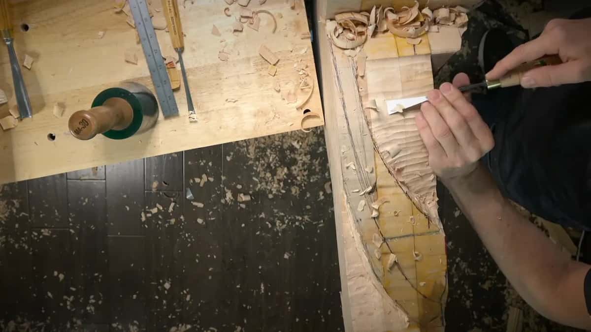 Both hands push a flat gouge along the top edge, shavings curling off in front of it