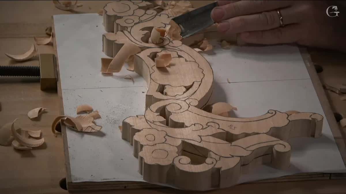 Gouge entering the sawn blank where the design is still only pencil lines, shavings gathering on the white board beneath