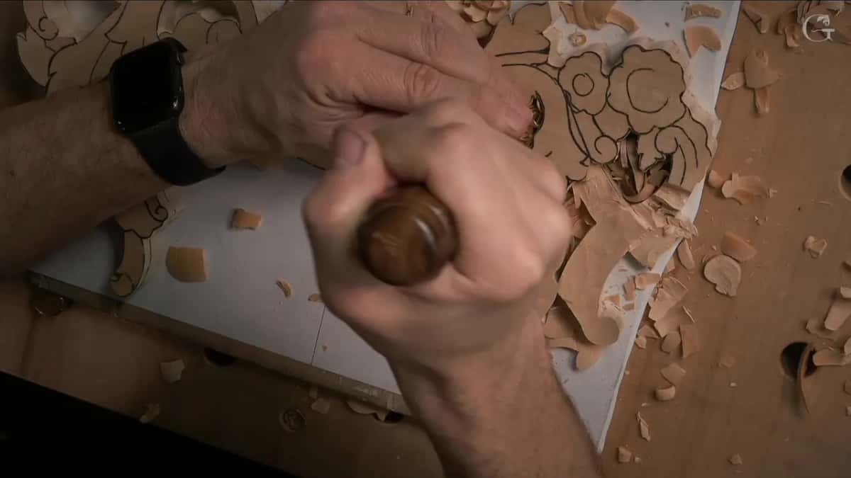 Both hands over the panel, one on the mallet and one steering the gouge through a flower