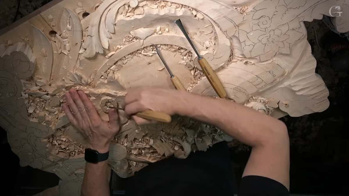 Overhead, both hands work at the left edge of the panel while two gouges lie on the carving