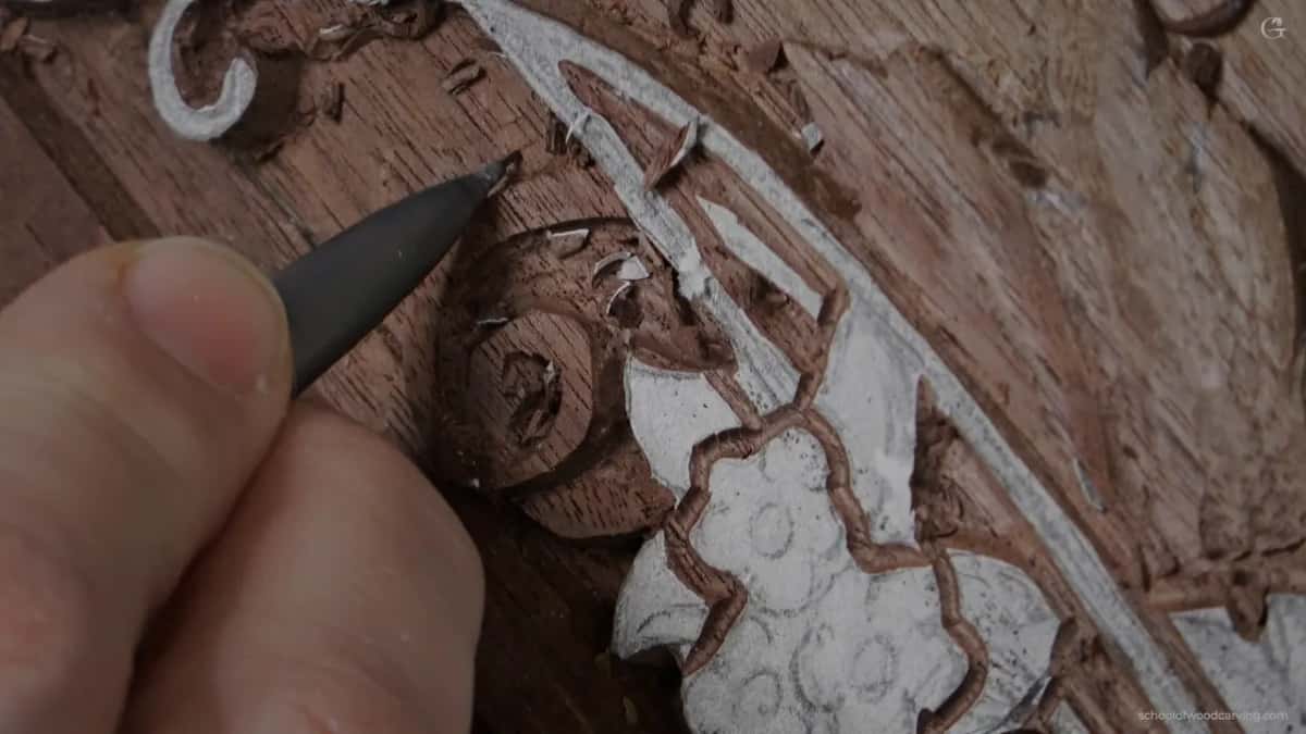 A wide dark blade held over the carving, thumb against the shank, about to take a whole area down at once.