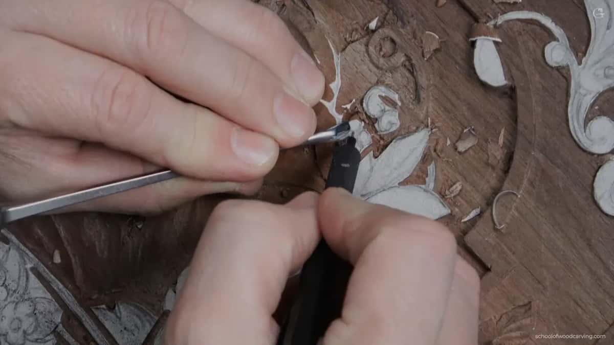 Both hands steady a fine gouge tipped over on the wood rather than held square to it.