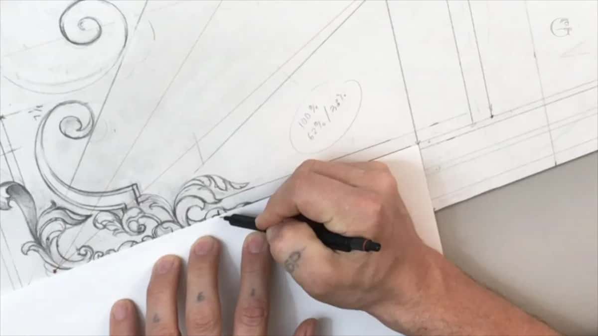 A hand marks the edge of a paper scrap laid along the bottom border, under the drawn scrolls