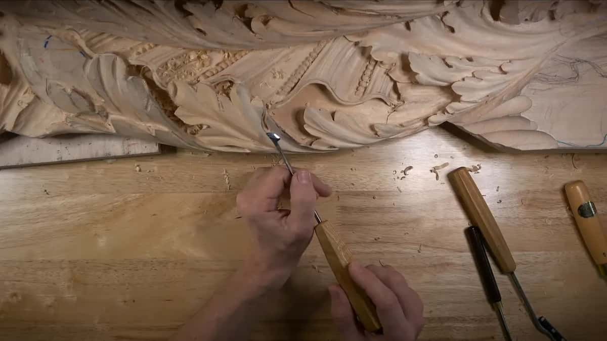 Underneath Both hands holding a small gouge up under the frieze, two more tools on the bench