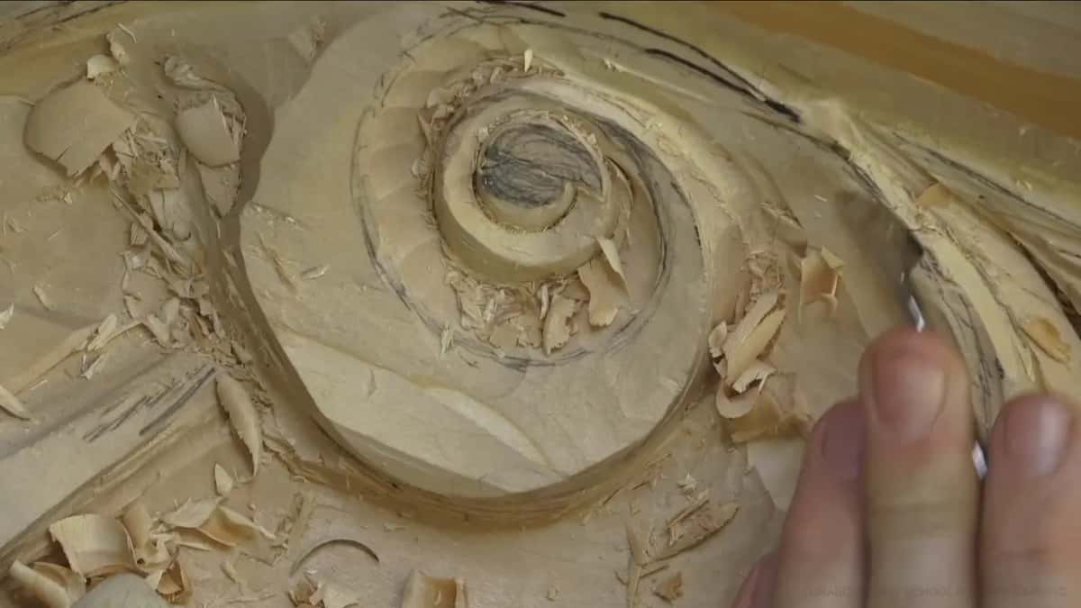Into the volute Spiral volute part carved, shavings everywhere, fingers holding a blade at the right of the frame