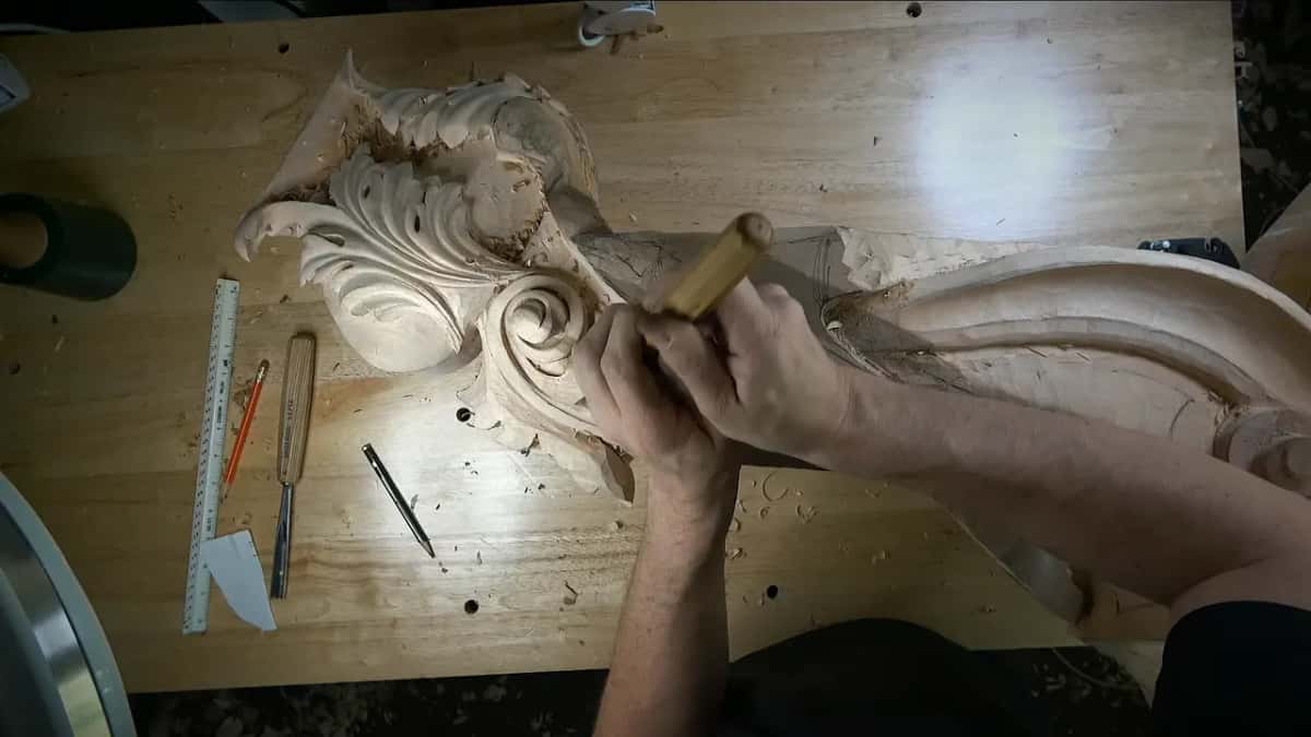 Two hands drive a gouge into the side below the scroll, chisels and a ruler set aside