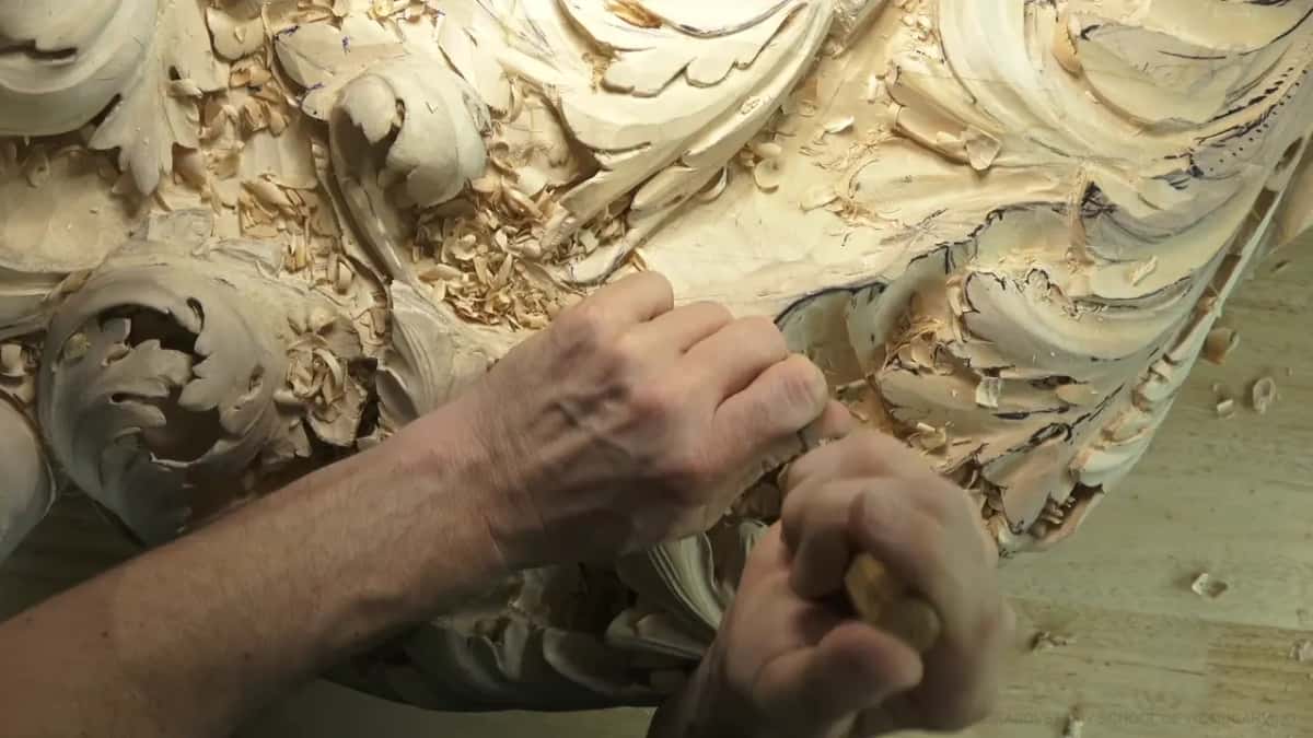 From above, both hands push a gouge into the curved rim of a carved shell, acanthus leaves all around.