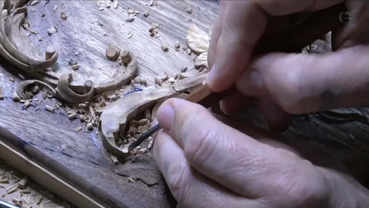 Both hands guide a small gouge into the fragile curl, now barely attached