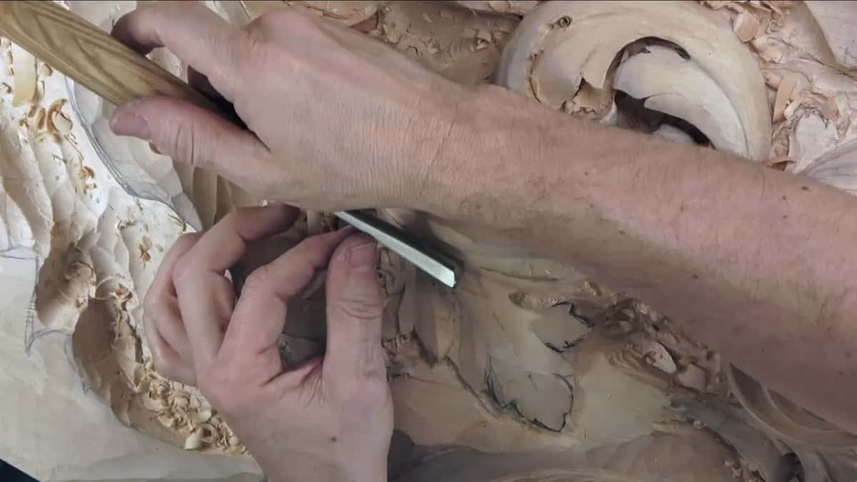 Two hands guide a long straight gouge along the rim of a curling acanthus leaf, shavings piled in its hollows.