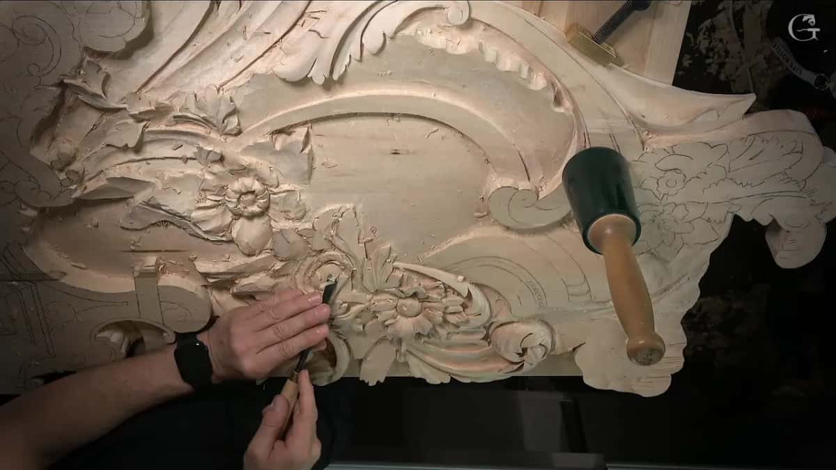 Overhead, both hands work the flower near the panel's edge while a mallet stands to the right