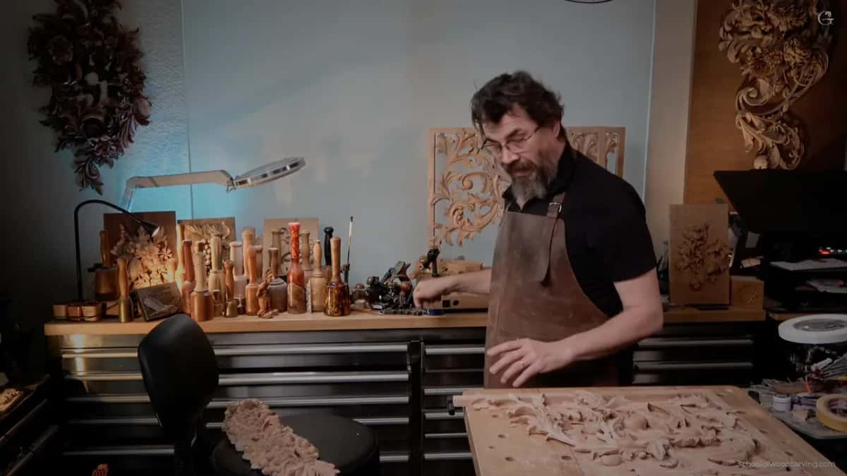 In a leather apron at his workbench, carved panels on the wall and rows of mallets behind him