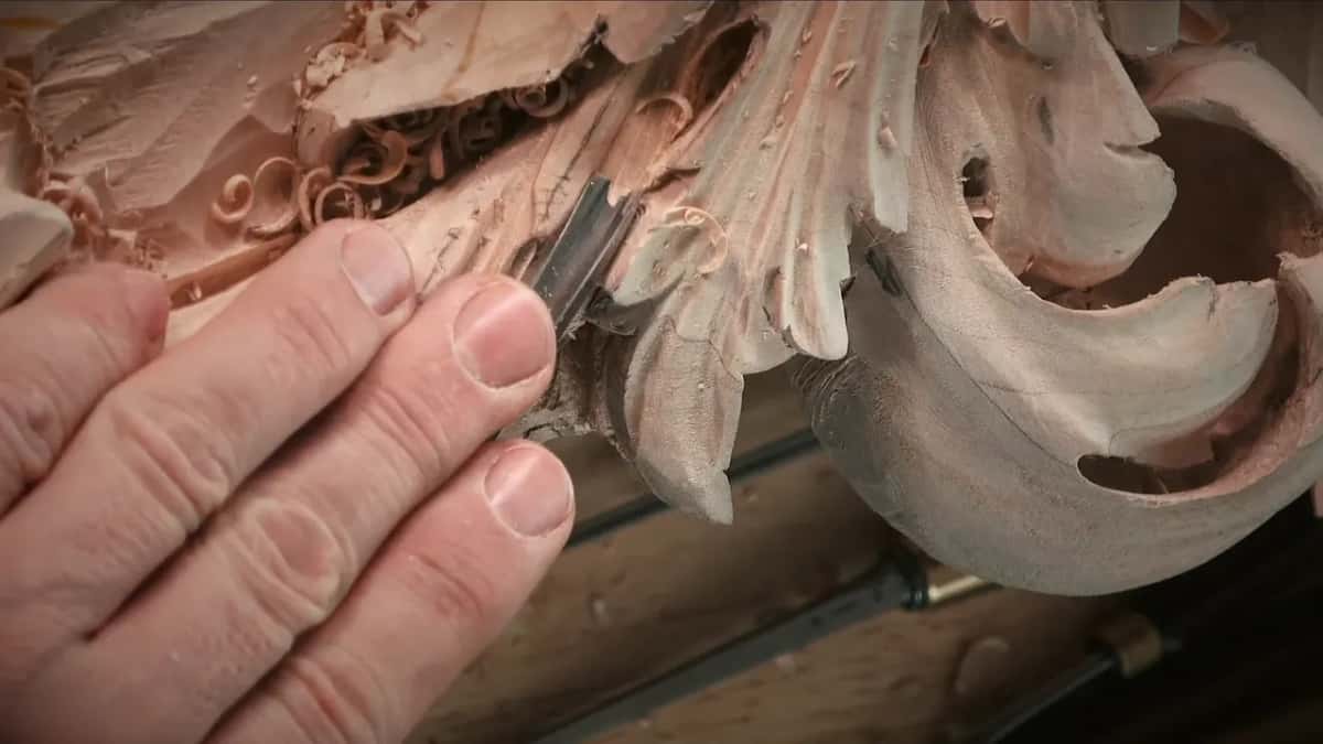Fingertips steering a small gouge along a raised lobe, curled shavings lying on the carving around it