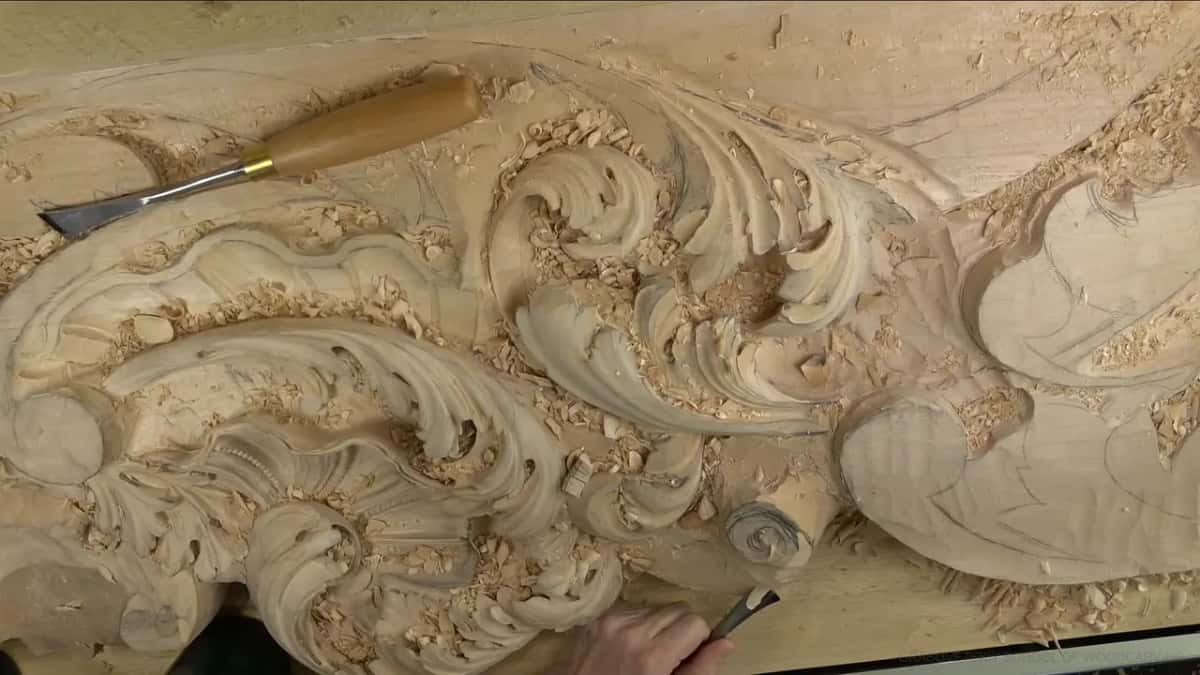 An orange-handled tool lies across the upper leaves while a hand works low on the board with a small gouge.